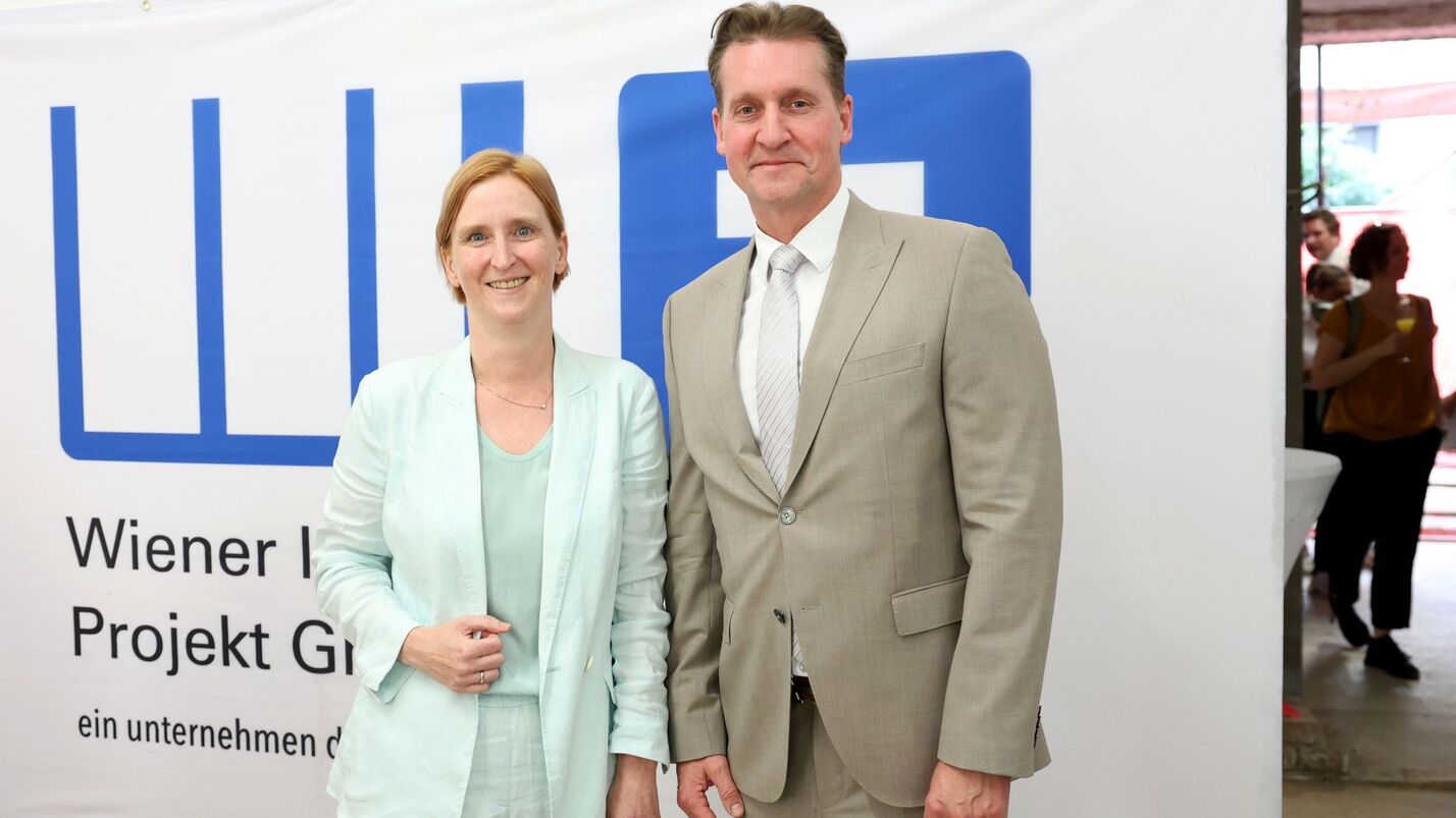 Zwei Personen in Anzügen stehen vor einem weißen Banner mit blauem Logo