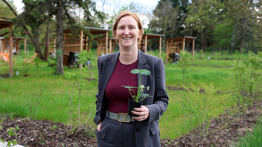 Person mit kurzem Haar und grauem Anzug hält einen kleinen Blumentopf mit einer Pflanze in einem Garten