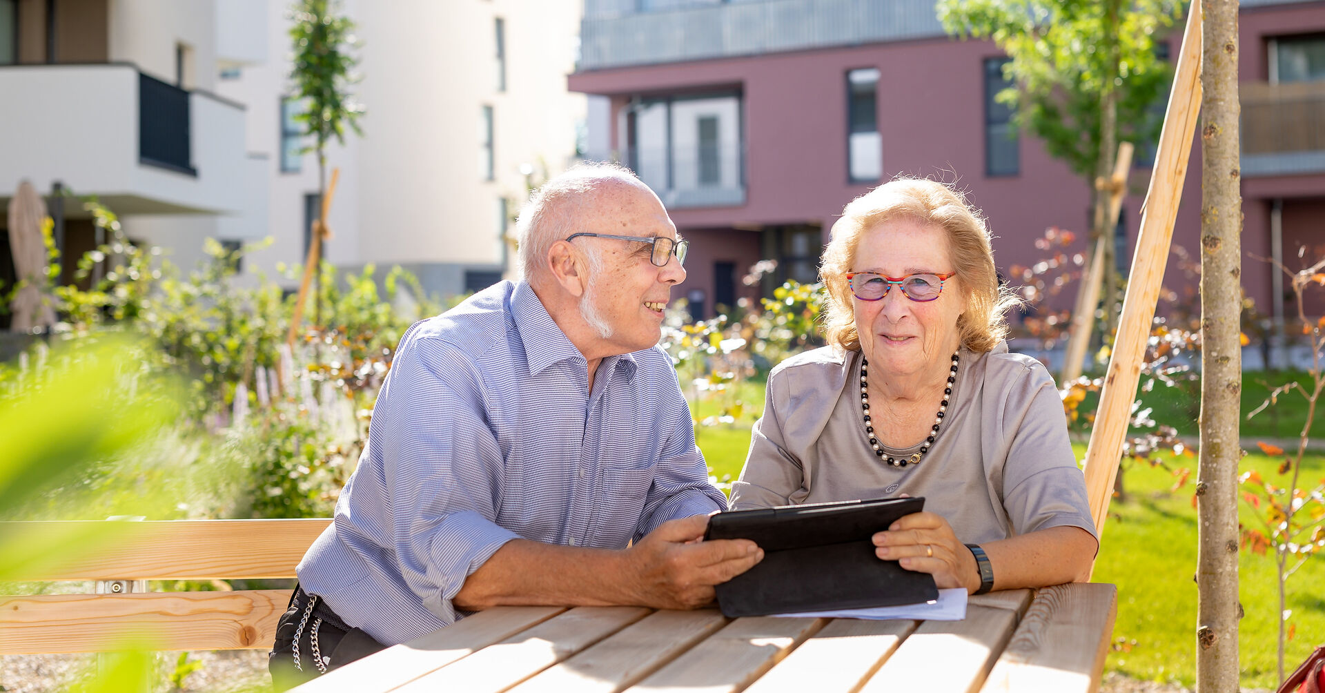 Ein älteres Pärchen, ein Mann und eine Frau, sitzen im Freien in einem Innenhof an einem Holztisch und schauen in ein Tablet.