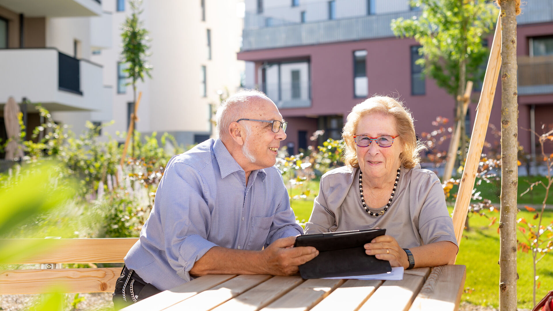 Ein älteres Pärchen, ein Mann und eine Frau, sitzen im Freien in einem Innenhof an einem Holztisch und schauen in ein Tablet.