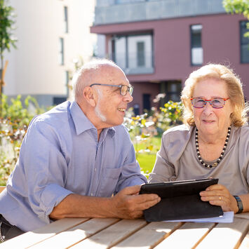 Ein älteres Pärchen, ein Mann und eine Frau, sitzen im Freien in einem Innenhof an einem Holztisch und schauen in ein Tablet.