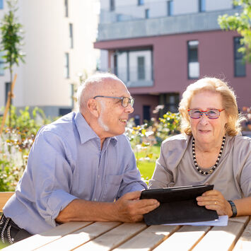 Ein älteres Pärchen, ein Mann und eine Frau, sitzen im Freien in einem Innenhof an einem Holztisch und schauen in ein Tablet.