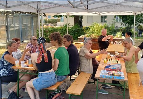 Mehrere Menschen sitzen an zwei Holzbänken unter einem weißen Pavillon und spielen Gesellschaftsspiele