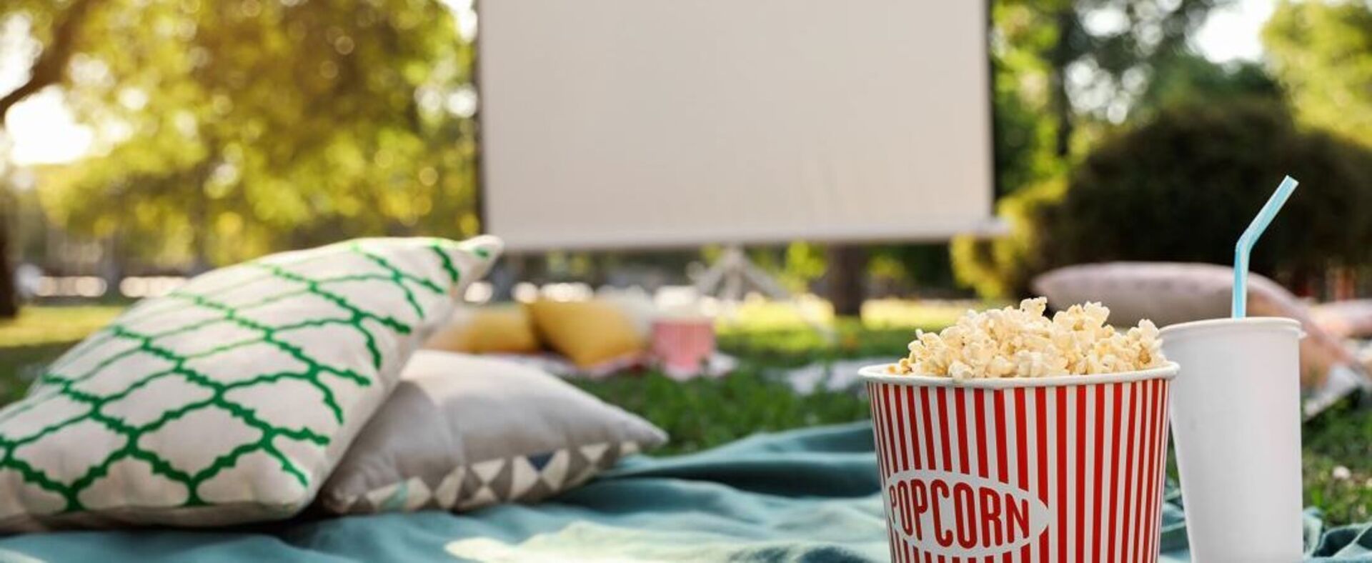 Sommerkino im Park: zu sehen sind eine Picknickdecke, Pölster, eine Popcorn-Tüte und ein Getränkebecher