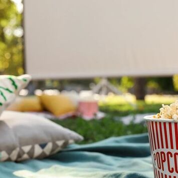 Sommerkino im Park: zu sehen sind eine Picknickdecke, Pölster, eine Popcorn-Tüte und ein Getränkebecher