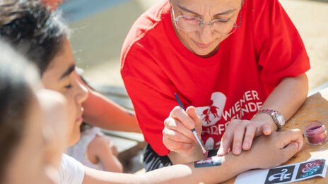 Frau in rotem T-Shirt malt mit Pinsel auf den Unterarm eines Kindes, mehrere Kinder sitzen um einen Tisch und halten Eis in der Hand.