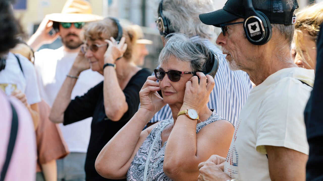 Mehrere ältere und jüngere Personen stehen im Freien und hören mit Kopfhörern aufmerksam zu, offenbar im Rahmen eines geführten Audiowalks.