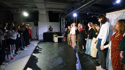 Rechts im Bild stehen Personen in einer Reihe und halten sich an den Händen, bereit zur Verbeugung, links eine Reiche von stehenden klatschenden Menschen. Der Raum ist mit punktuellen Scheinwerferlicht erhellt.