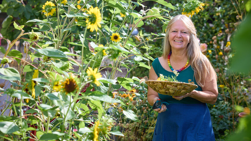 Frau mit langem Haar und bunter Halskette hält einen Korb mit Kräutern in einem Garten voller Sonnenblumen