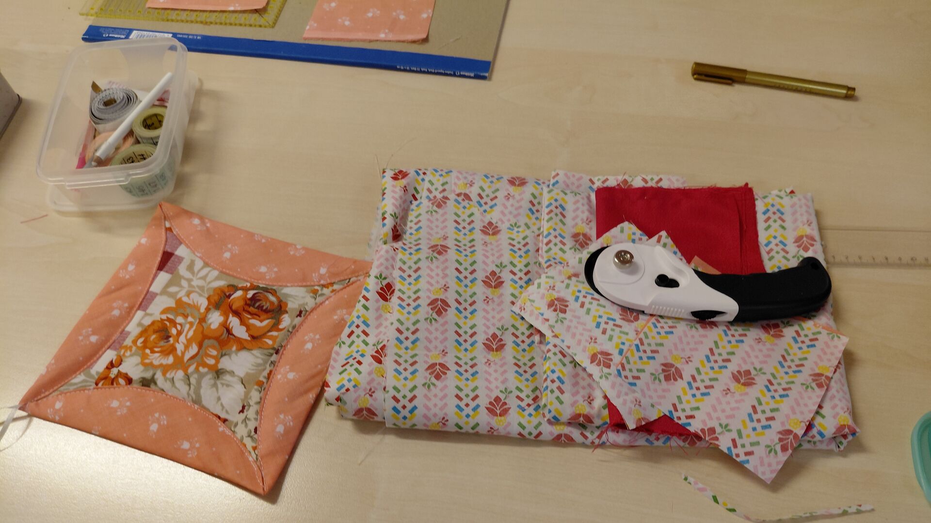 Gefalteter Stoff mit buntem Muster, darauf ein Rollschneider und ein rotes Stoffstück, daneben ein genähter Topflappen mit Blumenmuster und eine Box mit Nähzubehör auf einem Tisch