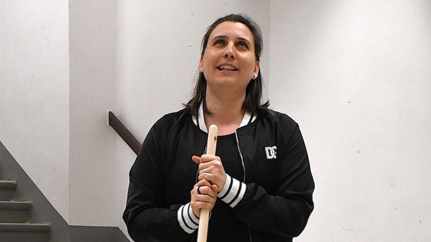 Eine Person steht mit einem Besen in der Hand und blickt freundlich lächelnd nach oben. Sie befindet sich in einem Raum mit Stiegenaufgang im Hintergrund.