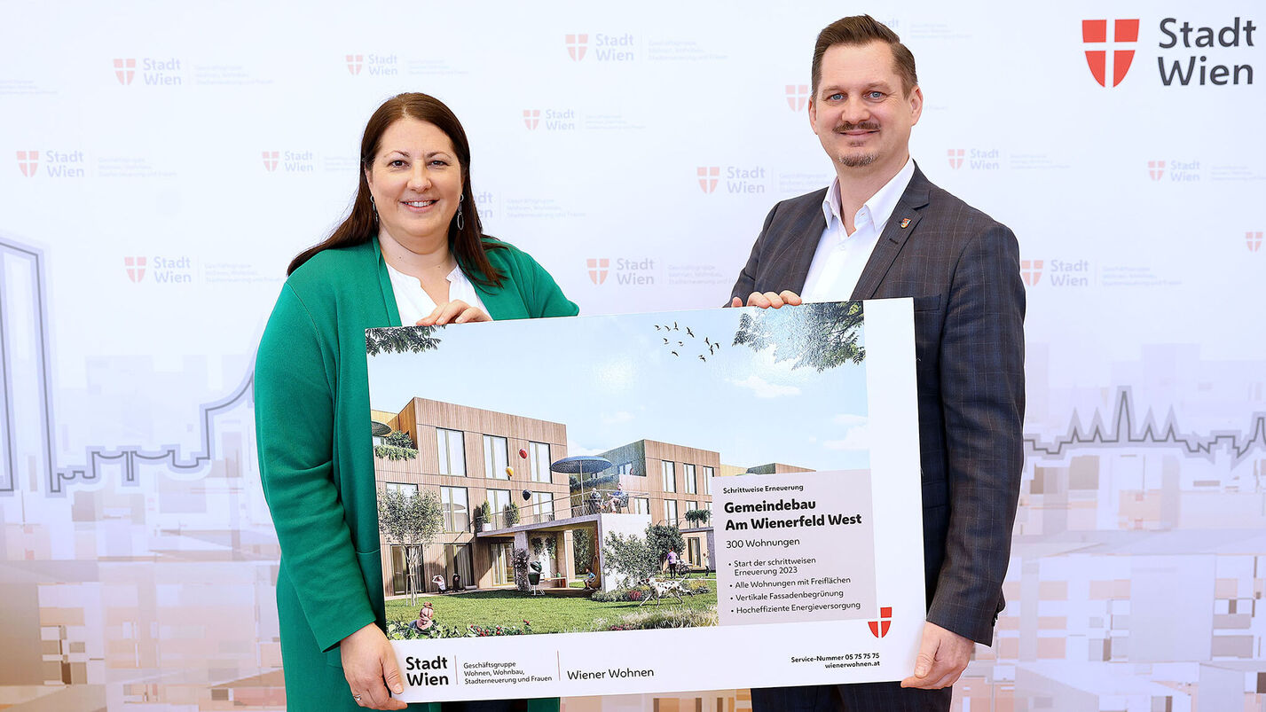 Eine Frau und ein Mann halten ein Schild mit der Visualisierung des neuen Gemeindebaus NEU in der Siedlung Wienerfeld West, erkennbar an der Beschriftung des Schildes. Sie lächeln in die Kamera.