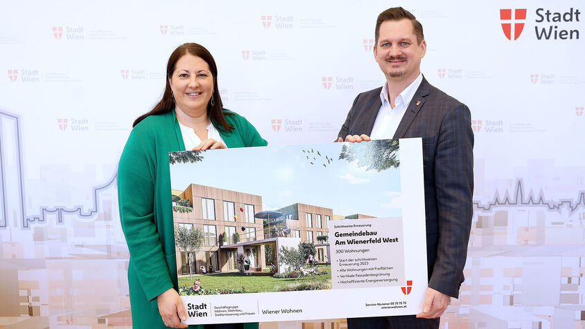 Eine Frau und ein Mann halten ein Schild mit der Visualisierung des neuen Gemeindebaus NEU in der Siedlung Wienerfeld West, erkennbar an der Beschriftung des Schildes. Sie lächeln in die Kamera.
