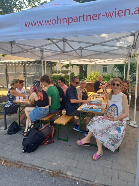 Gruppe von Menschen sitzt an zwei Holzbänken unter einem weißen Pavillon mit der Aufschrift 'www.wohnpartner-wien.at' und isst, trinkt und spielt Brettspiele im Freien