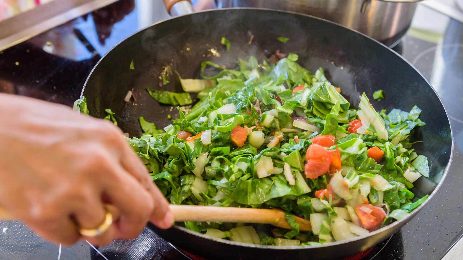 Eine Person rührt grünes Gemüse mit Tomaten in einer Pfanne auf dem Herd.