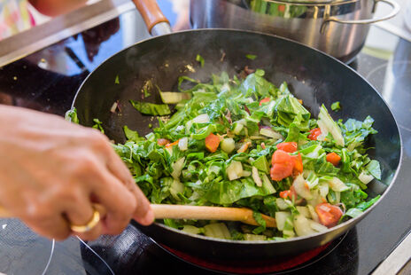 Eine Person rührt grünes Gemüse mit Tomaten in einer Pfanne auf dem Herd.