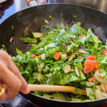 Eine Person rührt grünes Gemüse mit Tomaten in einer Pfanne auf dem Herd.
