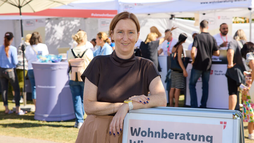 Frau lehnt an einem Schild mit der Aufschrift 'Wohnberatung Wien' auf einer Freiluftveranstaltung mit mehreren Menschen und Informationsständen im Hintergrund