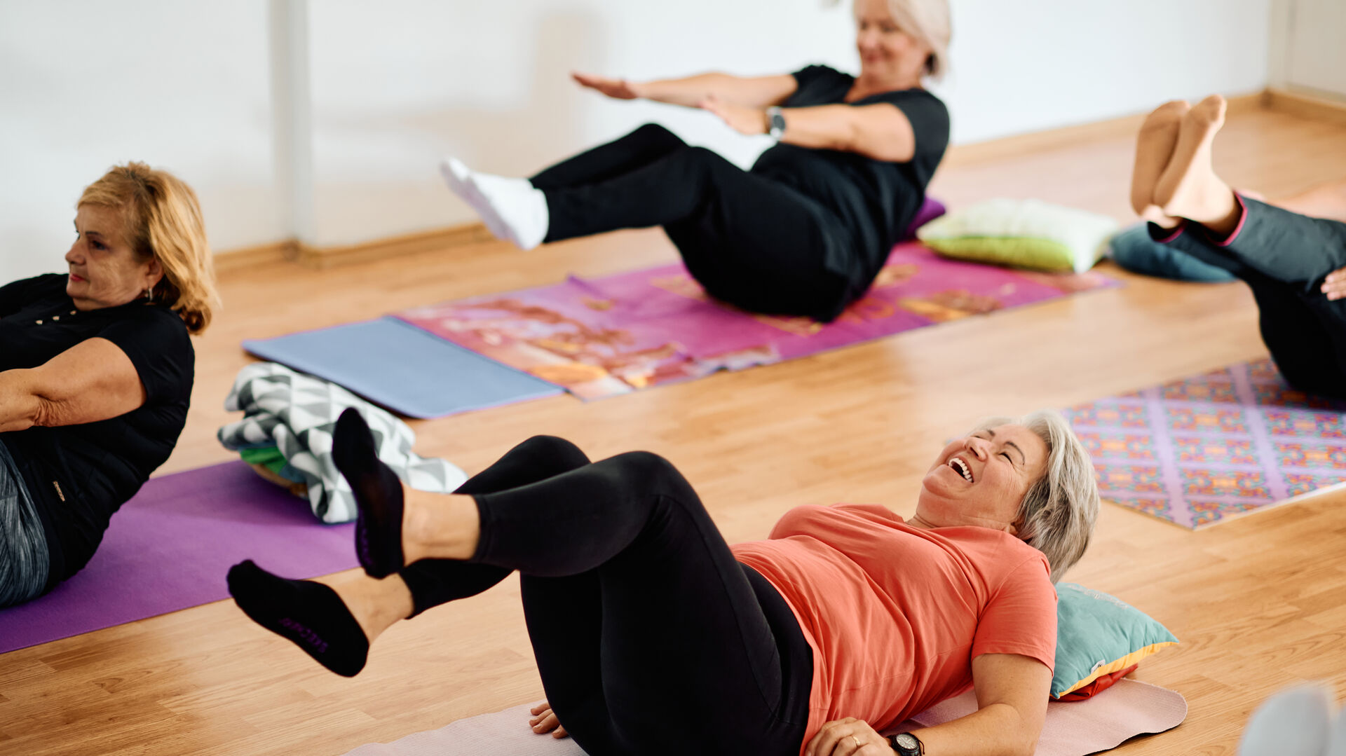 Ältere Damen liegen auf Gymnastikmatten am Rücken und machen Yogaübungen. Die Dame, die in der Mitte liegt, lacht.