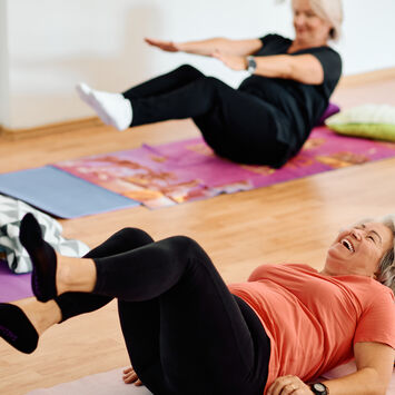 Ältere Damen liegen auf Gymnastikmatten am Rücken und machen Yogaübungen. Die Dame, die in der Mitte liegt, lacht.