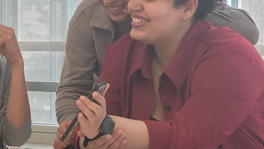 Zwei Frauen, ein sitzend, eine an ihr lehnend. Di sitzende zeigt ein Bild auf ihrem Handy, beide lächeln amüsiert