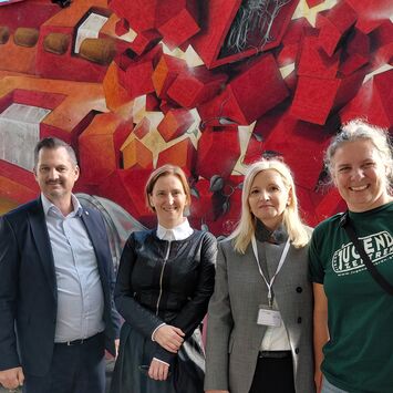 Vier Personen stehen vor einer bunten Wand mit rotem und orangefarbenem geometrischem Muster und lächeln in die Kamera