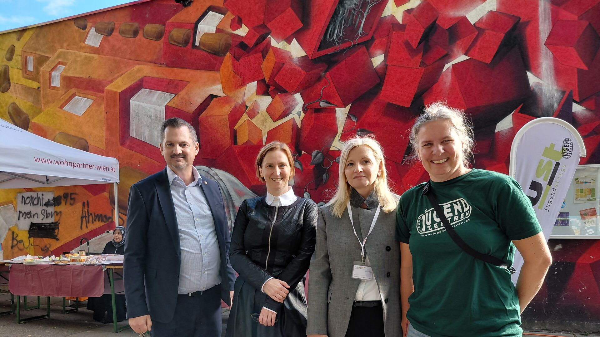 Vier Personen stehen vor einer bunten Wand mit rotem und orangefarbenem geometrischem Muster und lächeln in die Kamera