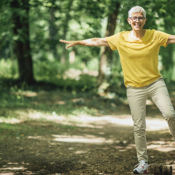 Eine ältere Frau balanciert im Wald in sportlich-sommerlicher Kleidung auf einem Baumstamm.