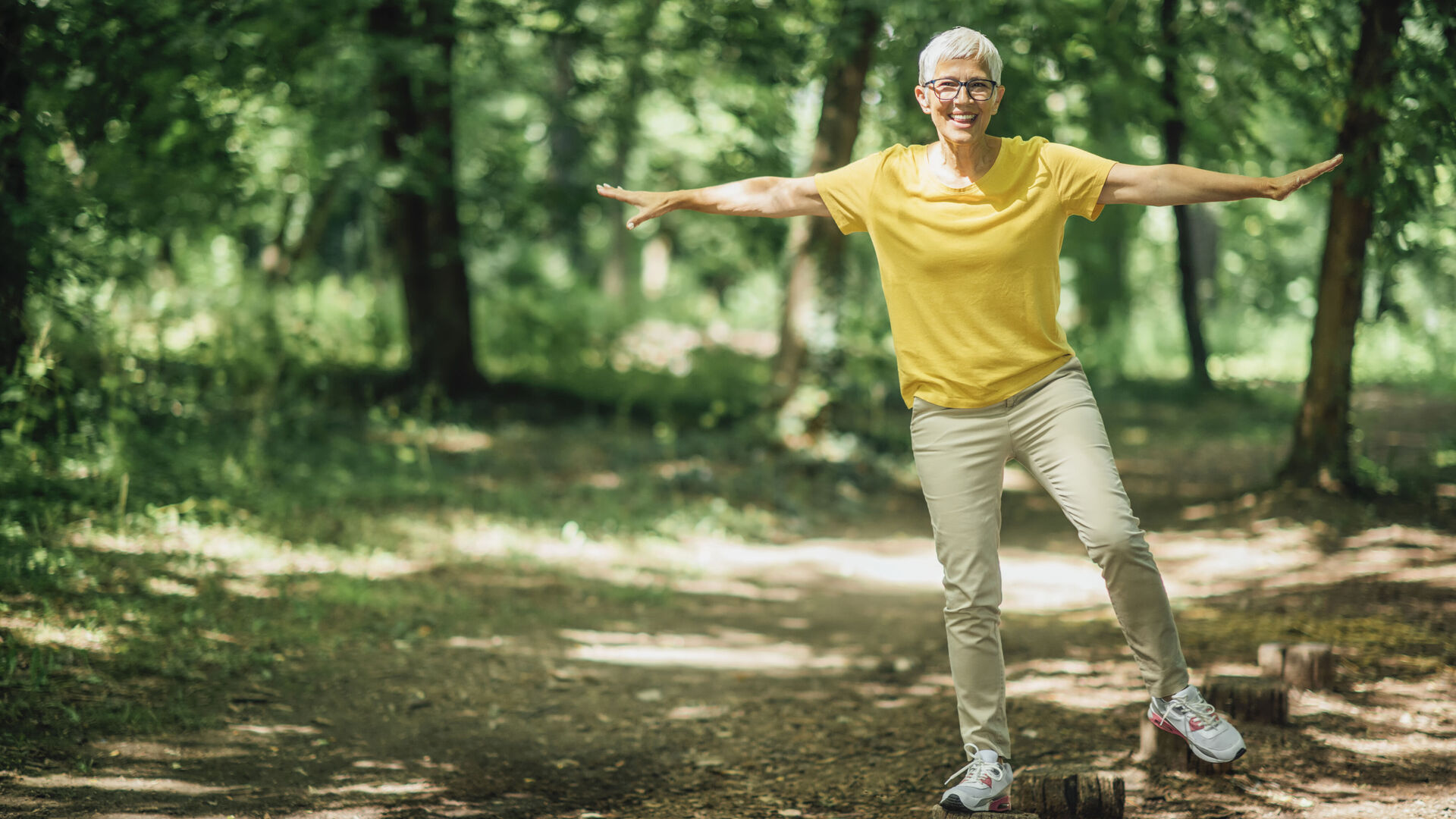 Eine ältere Frau balanciert im Wald in sportlich-sommerlicher Kleidung auf einem Baumstamm.