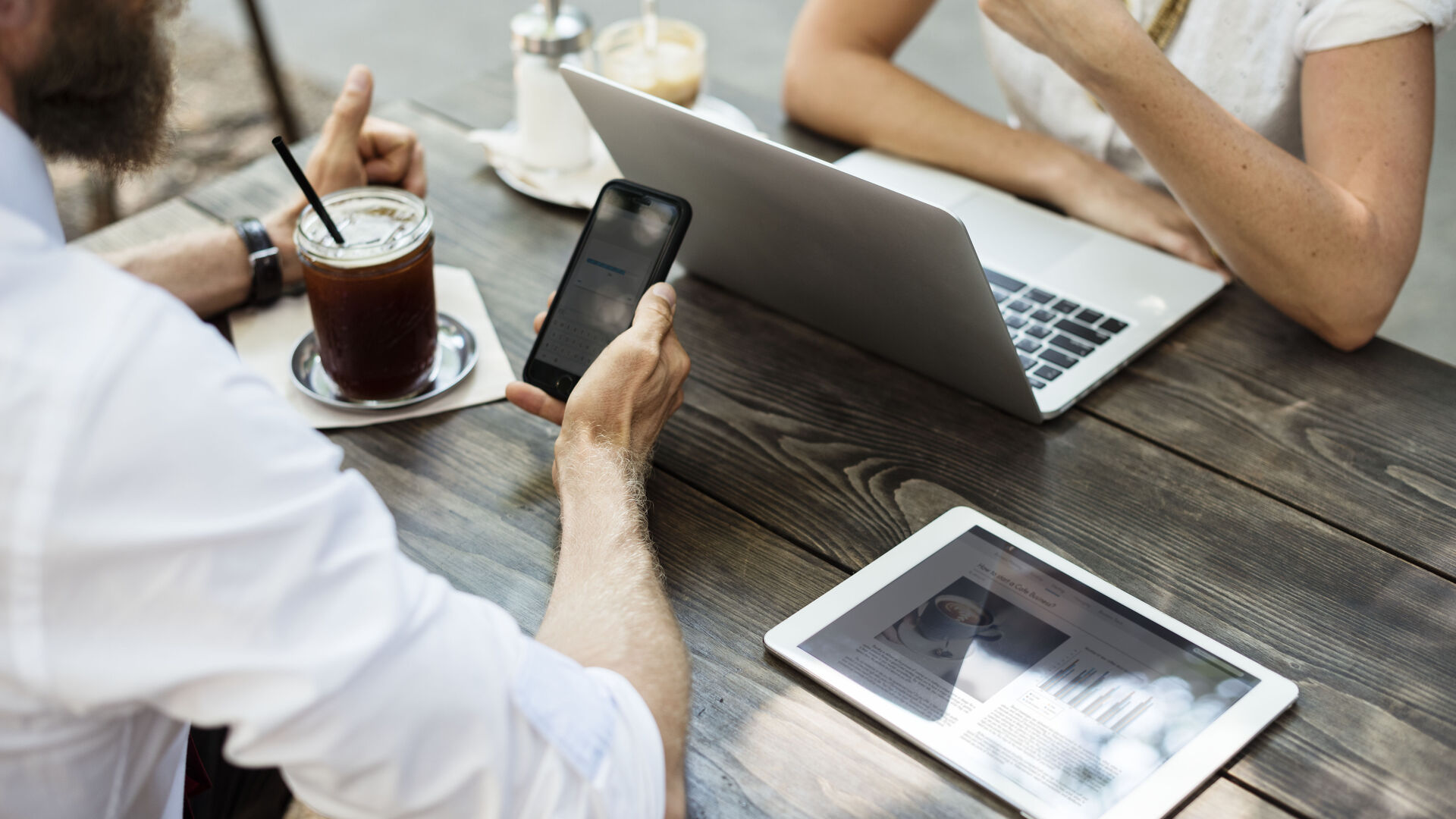 Zwei Leute sitzen an einem Holztisch im Freien. Der Mann hält ein Smartphone in der Hand und einen Kaffeebecher neben sich, die Frau sitzt vor einem Laptop. Ein Tablet liegt neben ihnen auf dem Tisch.