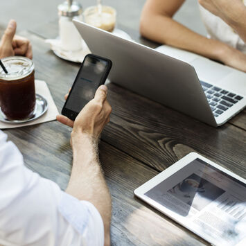 Zwei Leute sitzen an einem Holztisch im Freien. Der Mann hält ein Smartphone in der Hand und einen Kaffeebecher neben sich, die Frau sitzt vor einem Laptop. Ein Tablet liegt neben ihnen auf dem Tisch.