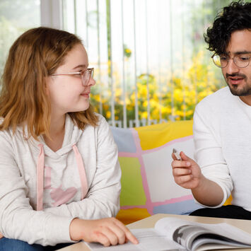 Ein Mädchen erhält Nachhilfe von einem etwas älteren Jungen. Die beiden sitzen an einem Tisch und schauen in ein Schulbuch.