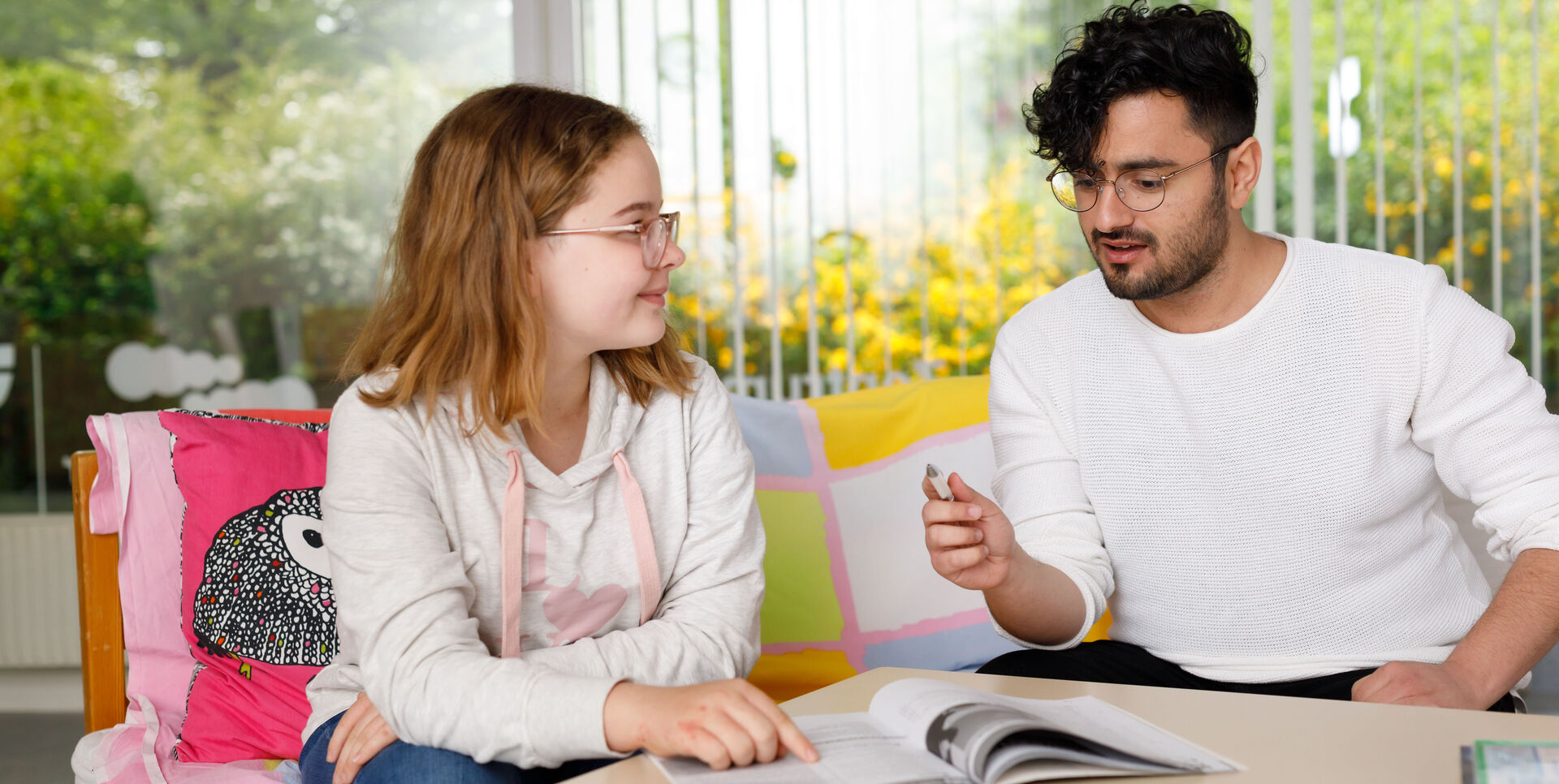 Ein Mädchen erhält Nachhilfe von einem etwas älteren Jungen. Die beiden sitzen an einem Tisch und schauen in ein Schulbuch.