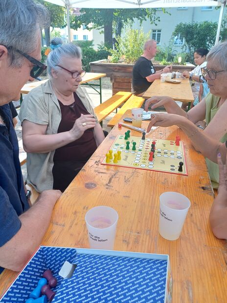 Drei ältere Personen sitzen an einem Holztisch im Freien und spielen ein Brettspiel mit bunten Spielfiguren