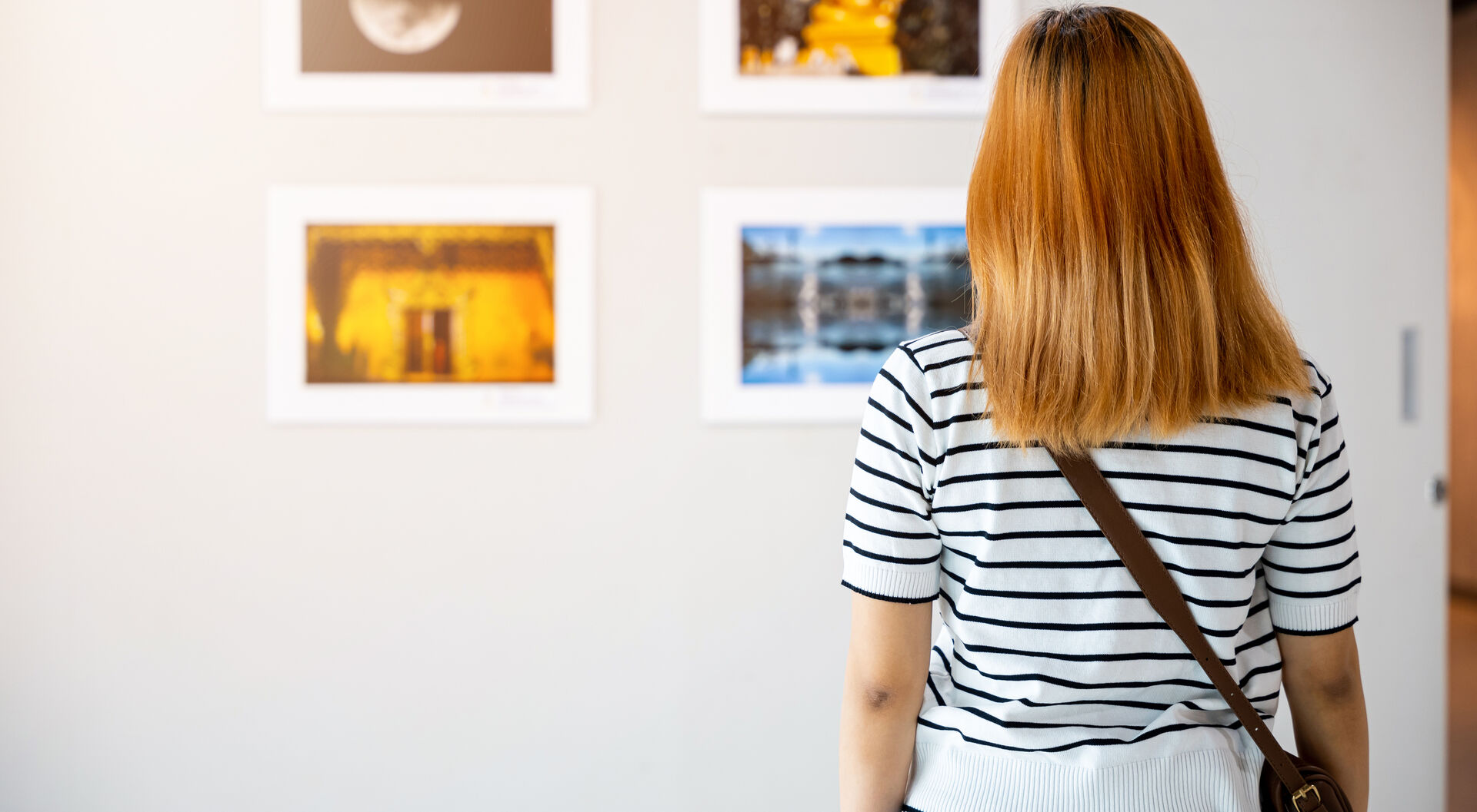 Frau mit gestreiftem Oberteil und Umhängetasche betrachtet vier gerahmte Bilder an weißer Wand