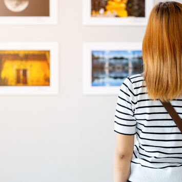 Frau mit gestreiftem Oberteil und Umhängetasche betrachtet vier gerahmte Bilder an weißer Wand