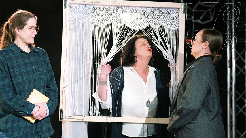 Drei PErsonen sind auf dem Bild zu sehen: eine blickt mit geschlossenen Augen und gespitzten roten Lippen aus einem Holrahmen mit Schnurvorhang der ein Fenster darstellen soll, eine andere steht davor und ist sichtlich irritiert von dem Anblick, die dritte steht daneben, hält ein Buch in der Hand und beobachtet die Szene.