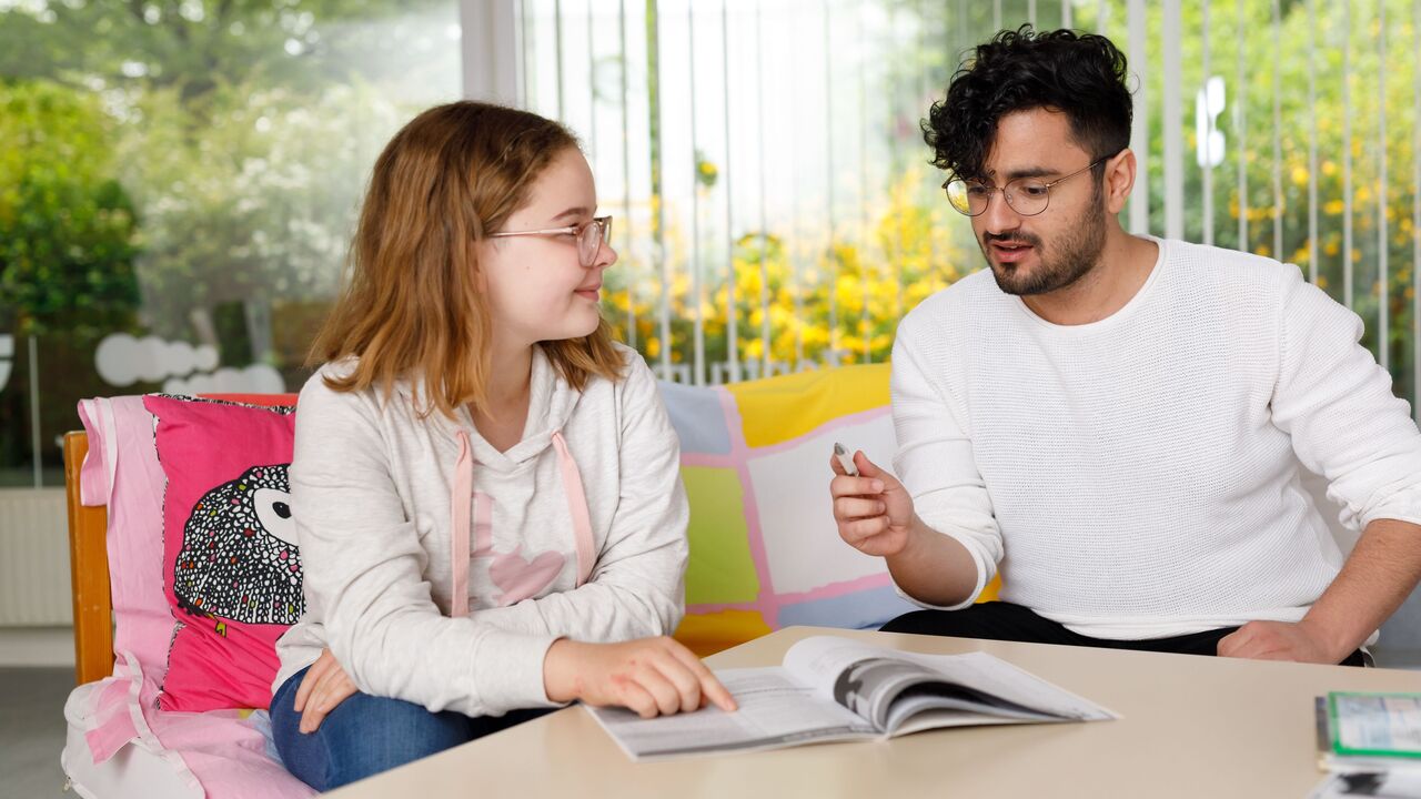 Ein junger Nachhilfelehrer gibt einem jungen Mädchen Nachhilfe, sie sitzen an einem Tisch über einem aufgeschlagenen Schulbuch.