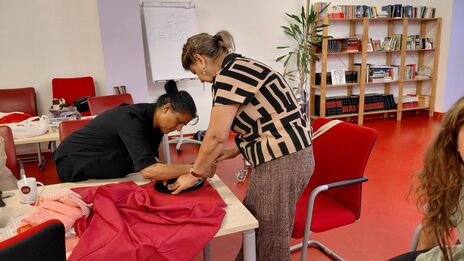 Zwei Frauen arbeiten gemeinsam an einem rot gepunkteten Stoff auf einem Tisch in einem Raum mit roten Stühlen und einem Bücherregal im Hintergrund