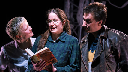 Drei Personen stehen nebeneinander, die mittlere hält ein offenes Buch in der Hand und spricht gerade. Die Person zu ihrer Linken blickt ihr direkt ins Gesicht, während die Person zur Rechten in das Buch blickt.