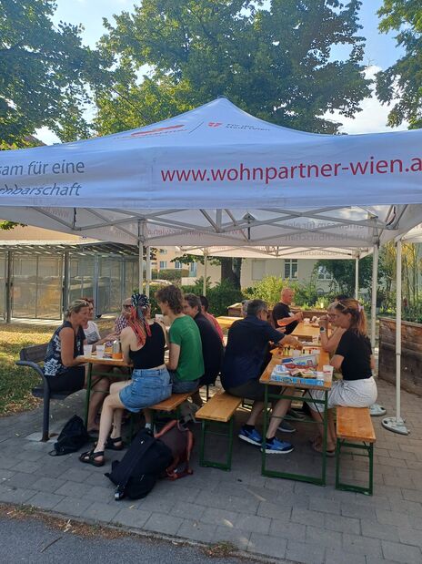 Gruppe von Menschen sitzt an zwei Holzbänken unter einem weißen Pavillon mit der Aufschrift 'www.wohnpartner-wien.at' und isst und trinkt im Freien