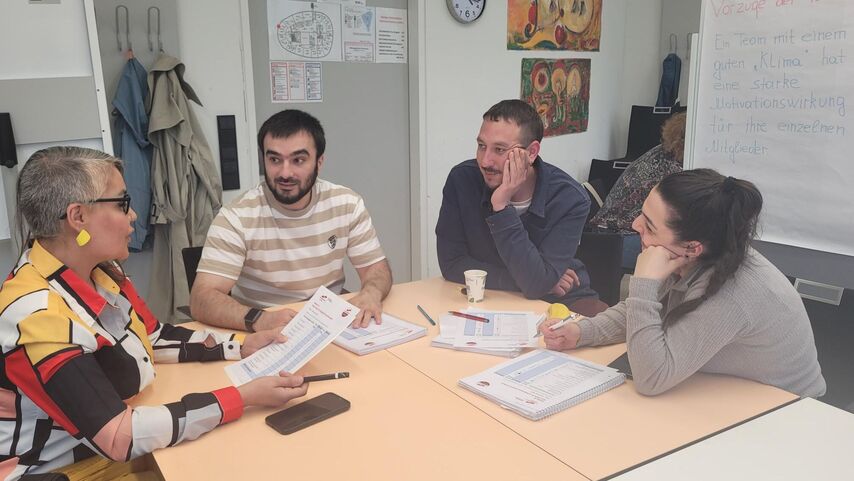 Vier Personen an einem Tisch sitzend. Drei Personen lauschen offenbar den Erzählungen einer Frau