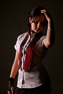 Eine Person mit kurzärmeliger weißer Bluse und roter Krawatte leger um den Hals geschlungen hält sich die linke Hand an den Mittelscheitel. Sie trägt einen silbernen Armreif am linken Arm und einen schwarzen Fischnet-Fingerling auf der rechten Hand, die am Körper herunterhängt.