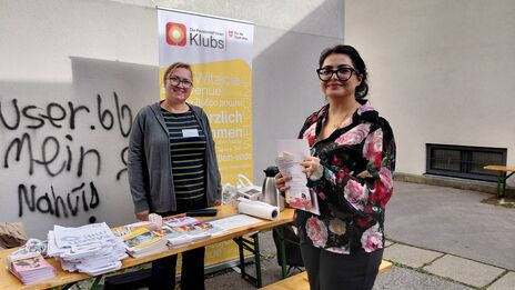 Zwei Frauen an einem Infostand mit Flyern und Broschüren vor einer Graffiti-Wand und einem Roll-Up der Pensionist*innen-Klubs