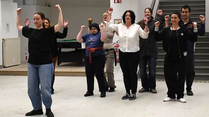 Einige Personen stehen in einem schmucklosen Raum mit Treppe im Hintergrund in zwei Reihen und sind von vorne zu sehen. Eine Person steht davor, alle haben die Arme nach oben abgewinkelt und die Hände zur Faust geballt.. 