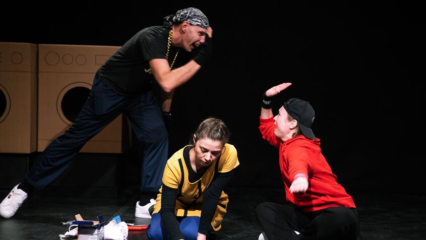 Szenenbild aus dem Gemeindebautheater: Ein schwarz gekleideter Jugendlicher mit Kopftuch gibt einer auf dem Boden sitzenden Jugendlichen mit rotem Sweater und verkehrt getragenem schwarzen Baseballcap High Five. Dazwischen kniet eine Jugendlich mit gelbem Oberteil und zeichnet mit Kreide etwas auf den Boden.