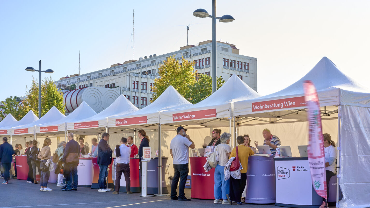 Menschen stehen an mehreren weißen Zelten mit roten Bannern und Informationsständen in einer sonnigen Straße vor einem großen Gebäude