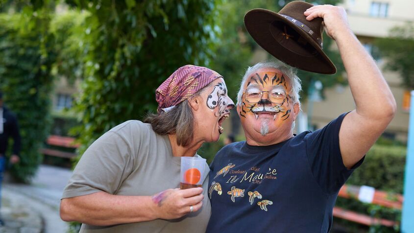 Eine Frau und ein Mann sind wie ein Hund und ein Tiger geschminkt, der Hund (die Frau) bellt offenbar den Tiger an, der freundlich in die Kamera lächelt und dabei seinen Hut zieht.