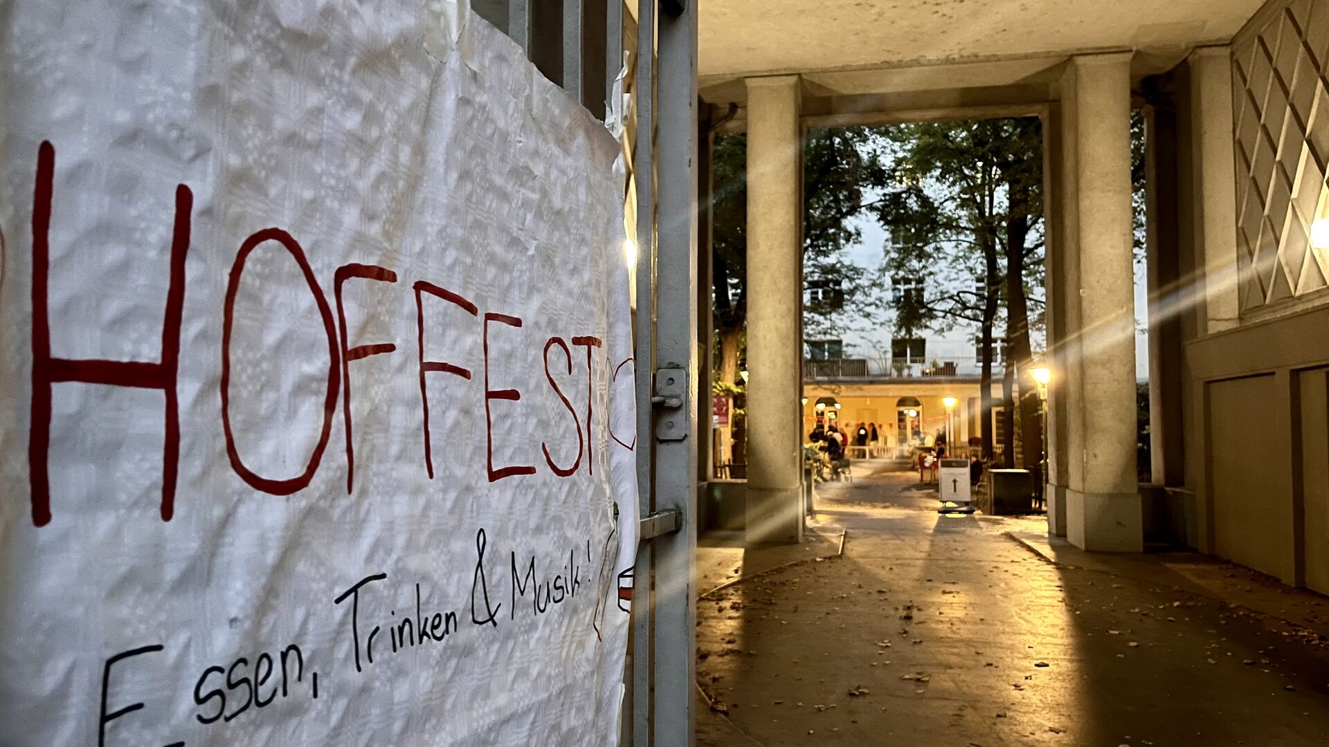 Handgeschriebene Plakatankündigung für ein Hoffest mit Essen, Trinken und Musik an einem Tor mit Blick auf einen beleuchteten Innenhof