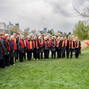 Der 1. Wiener Gemeindebauchor steht mit Chorleiterin Nelli Goncharuk auf einer Wiese und probt.